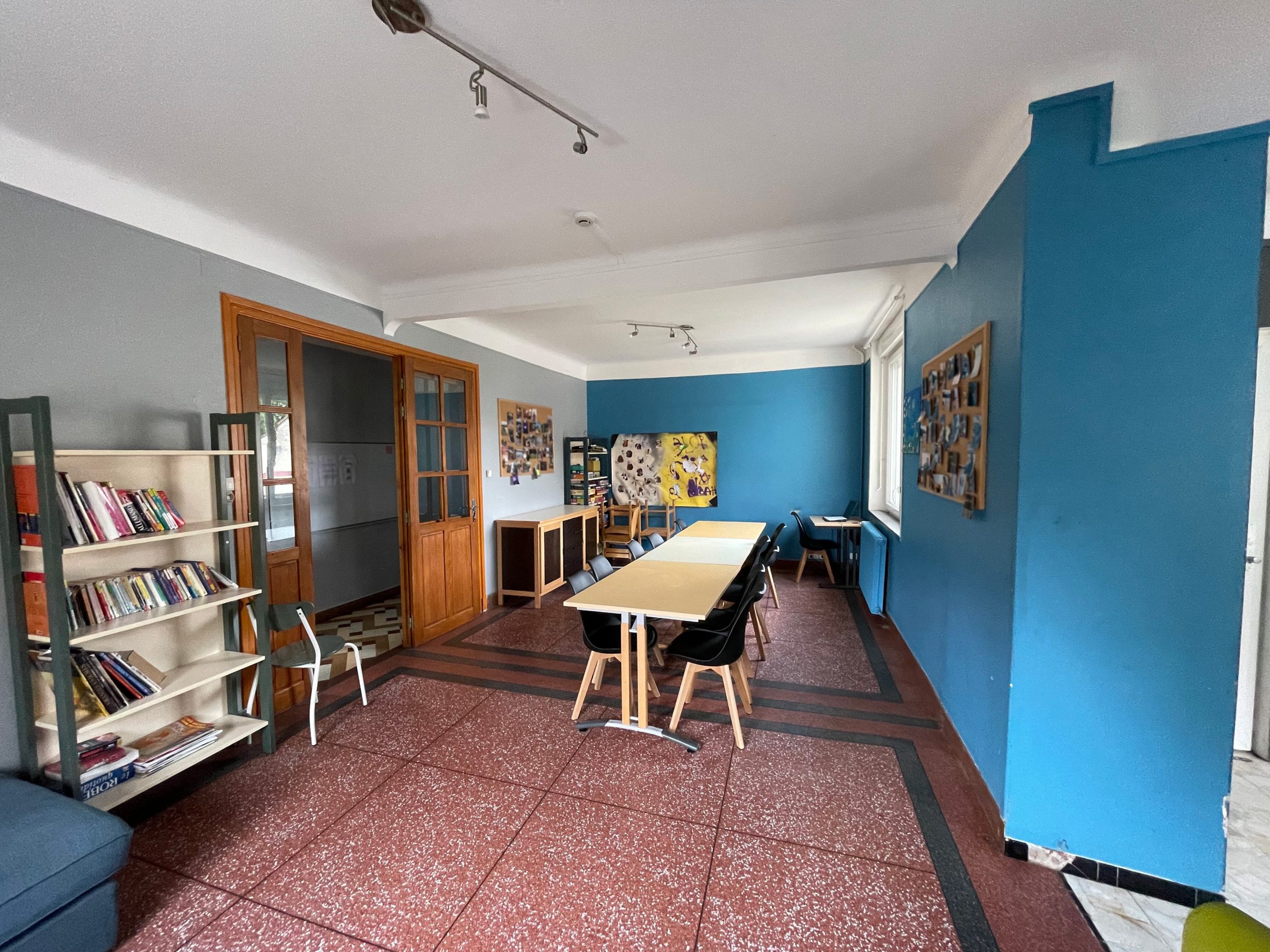 Salle de vie commune de la MECS de l'Allée Neuve montrant une grande table, des chaises, des étagères dont une remplie de livre et l'autre de jeux de sociètes