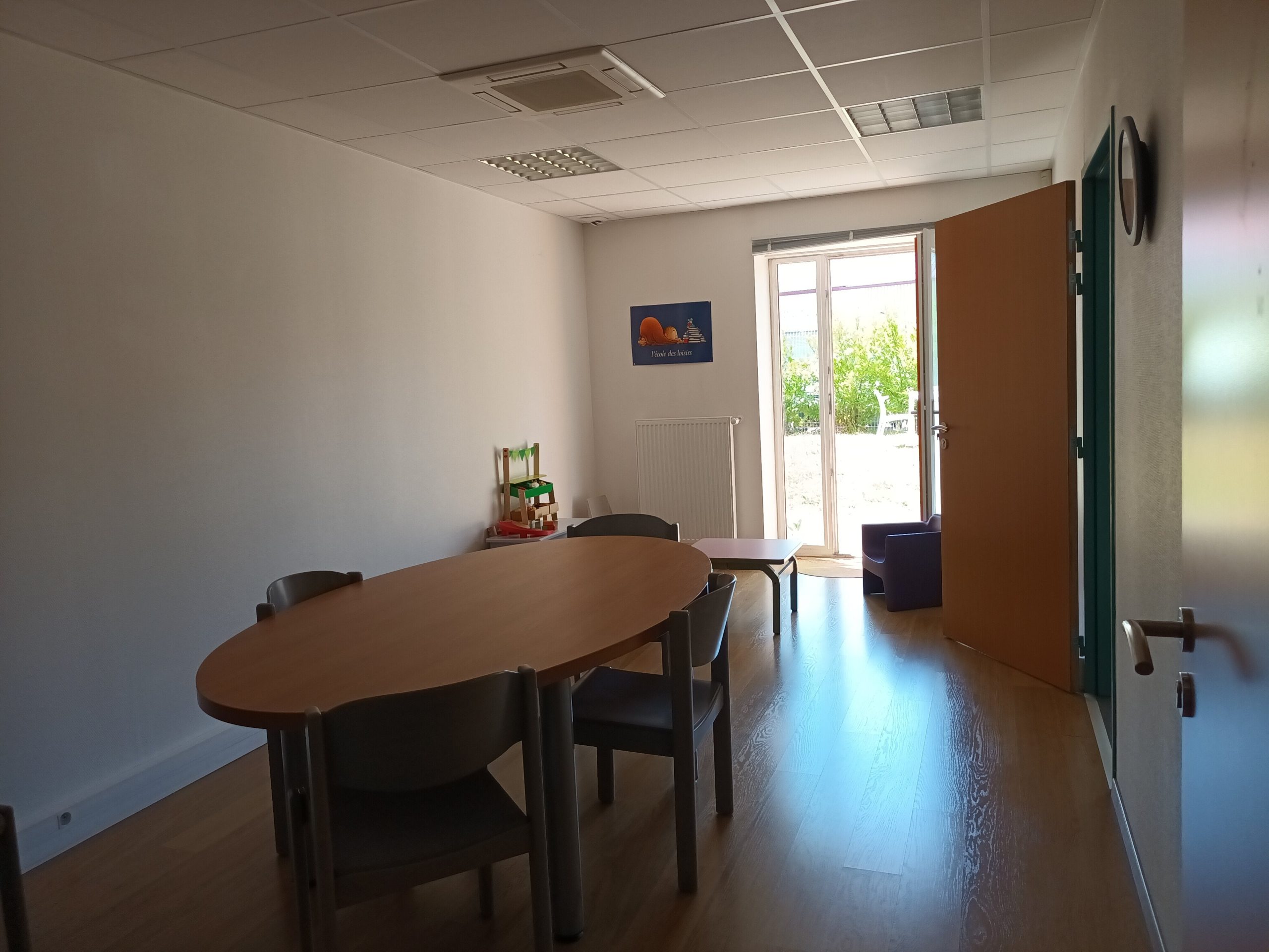 Salle de rencontre du SRP REALISE, montrant une table ovale avec des chaises, des jouets pour enfants en bas âge, avec une fenêtre montrant un extérieur verdoyant